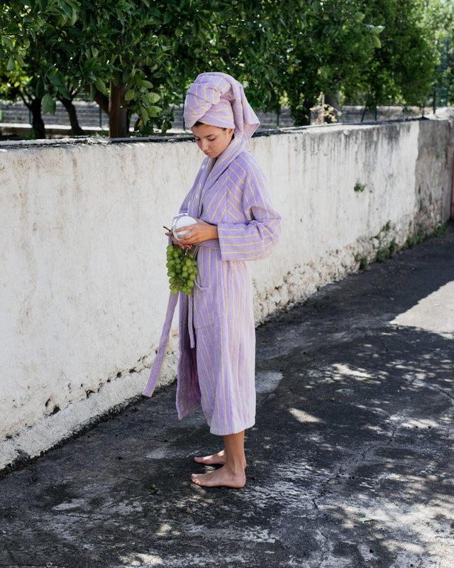 Frau im Bademantel NARAM Lilac & Neon Yellow mit Handtuch-Turban vor einer weißen Mauer, Trauben in der Hand