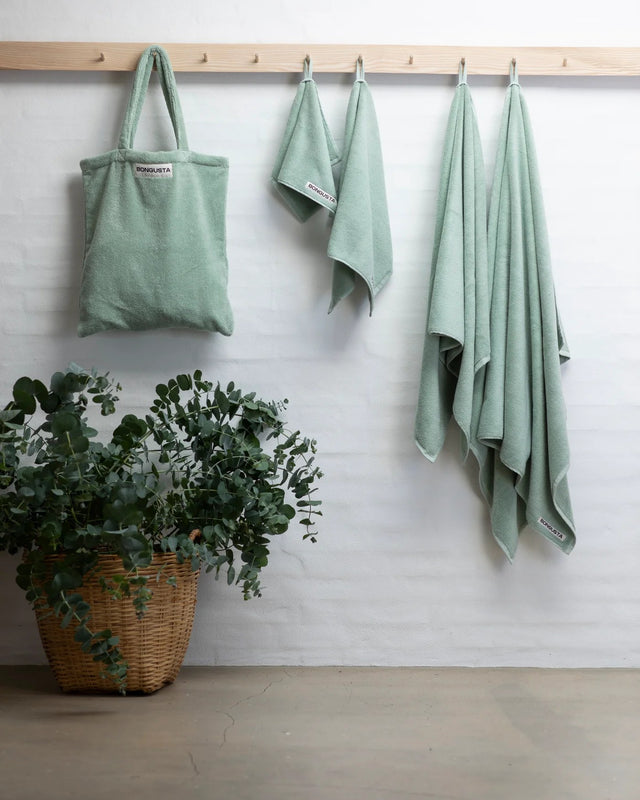 Tote Bag in Silver Sage an einer Hakenleiste aus Holz neben Handtüchern in derselben Farbe, davor ein Korb mit Eukalyptus
