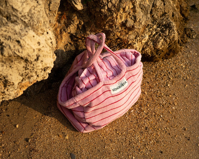 Rosa gestreifte Weekend-Bag NARAM im Abendlicht am Sandstrand, an einen Felsen gelehnt, darin ein Frottee-Handtuch