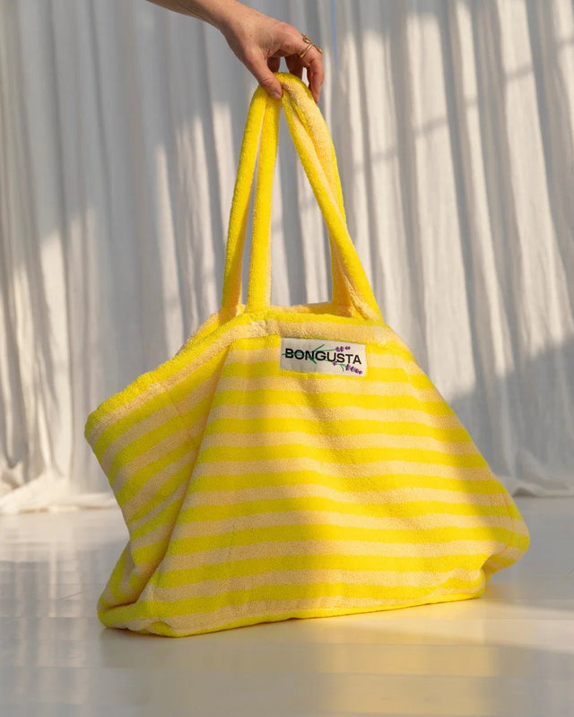Hand hält die Weekend-Bag Pristine & Neon Yellow an den Henkeln vor einem weißen Vorhang im Sonnenlicht