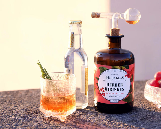 Herber Hibiskus mit Tonic im Tumbler mit Rosmarinzweig, daneben Flasche mit Ausgießer und Tonic in der Abendsonne