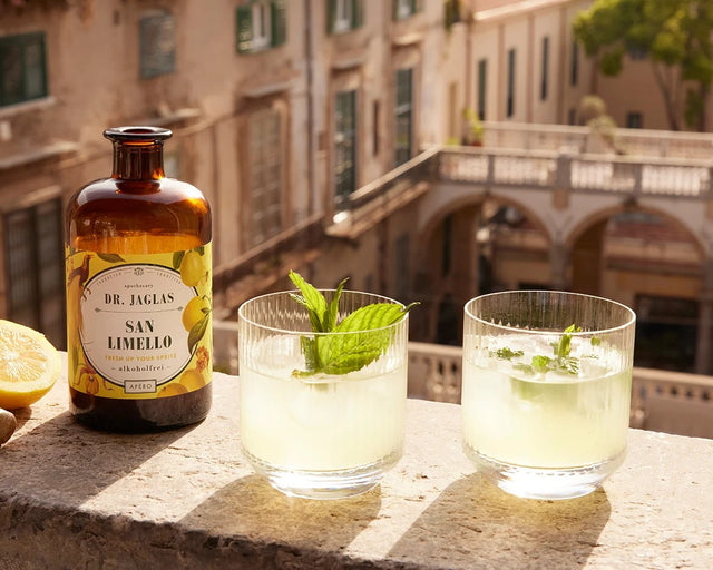 Zwei Tumbler San Limello mit frischer Minze auf einer Steinmauer, daneben Flasche und halbe Zitrone
