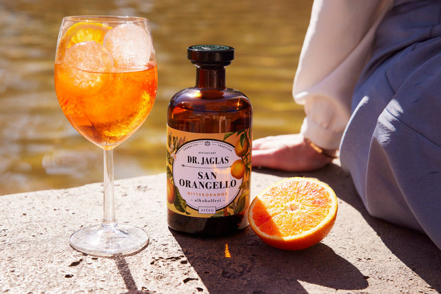 Orangello Spritz im Weinglas mit Eiswürfeln und Orangenscheibe neben der Flasche San Orangello und einer halben Orange