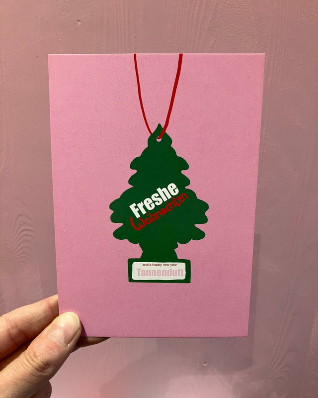 Weihnachtspostkarte „Freshe Weihnachten“ von hej ibiza: grüner Duftbaum an roter Schnur mit „Tannenduft“-Etikett auf Rosa, in der Hand vor einer lila Holzwand
