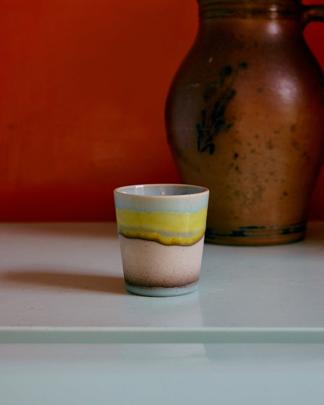 Kaffeebecher POP ART in glint mit gelbem Streifen zwischen Hellblau und Sand, vor roter Wand neben einem alten Steingutkrug