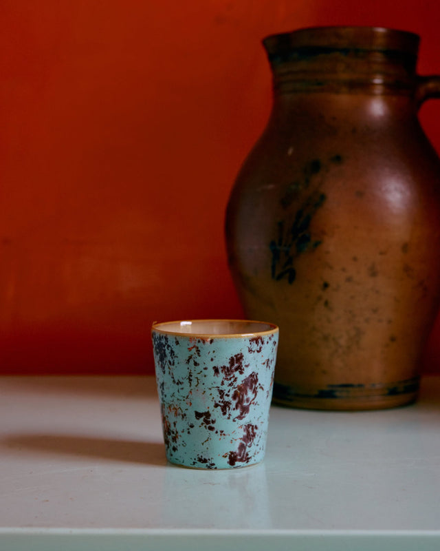 Kaffeebecher SURREAL in patina, vor roter Wand neben einem alten Steingutkrug