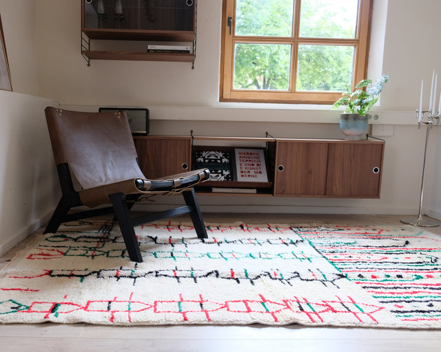 Berberteppich HOUSES 1,40 x 2,50 m im Raum: Reihen kleiner Häuser in Rot, Grün und Schwarz, daneben ein Ledersessel