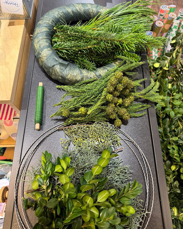 Material für den Adventskranz-Workshop auf dem Tisch im Laden: Kranzrohling, verschiedenes Tannengrün, Buchsbaum und grüner Wickeldraht.