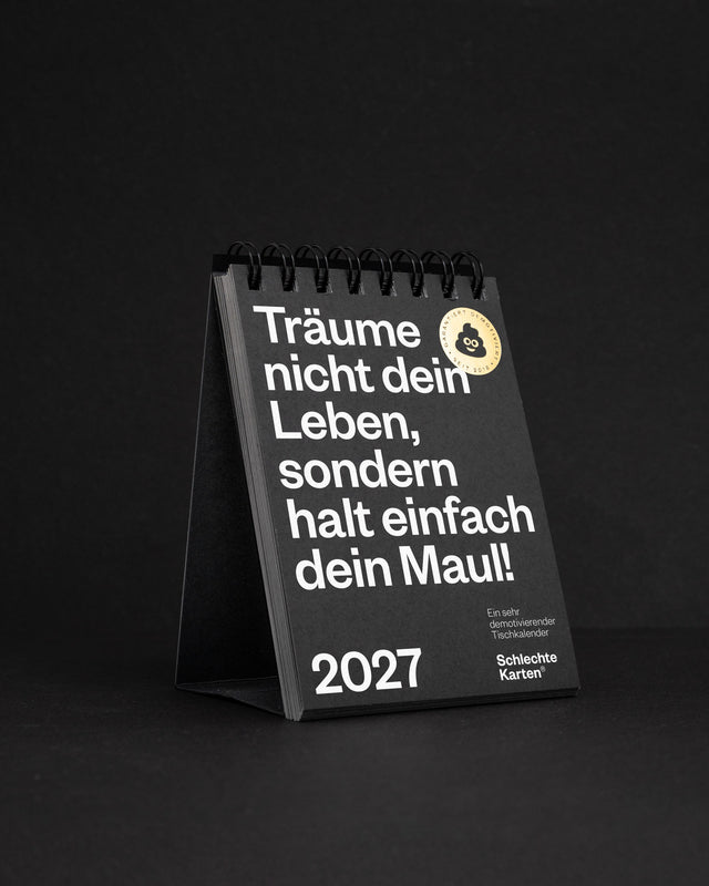 Titelseite des Tischkalenders 2027 mit goldrundem Aufkleber, fotografiert auf schwarzem Hintergrund.