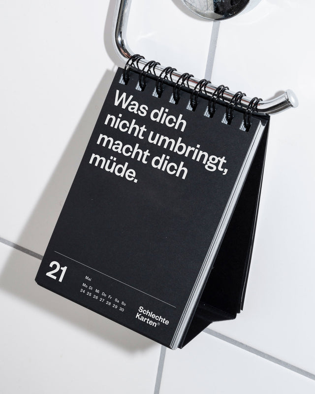 Der Tischkalender hängt an einem verchromten Handtuchhalter über weißen Badezimmerfliesen.
