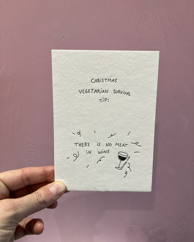 Letterpress-Weihnachtskarte von Studio Flash: schwarze Handschrift „Christmas vegetarian survival tip: there is no meat in wine“ mit kleinem Rotweinglas und Luftschlangen auf weißem Baumwollpapier, in der Hand vor einer rosa Holzwand