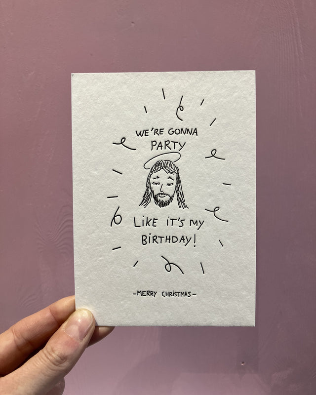 Letterpress-Weihnachtskarte von Studio Flash: gezeichneter Jesus mit Heiligenschein zwischen Luftschlangen, darum „We’re gonna party like it’s my birthday!“ und unten „Merry Christmas“, in der Hand vor einer rosa Holzwand