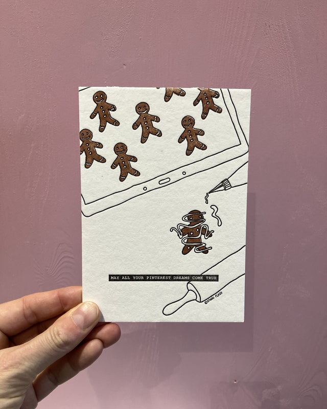 Letterpress-Weihnachtskarte von Studio Flash: perfekte braune Lebkuchenmännchen auf einem Tablet, darunter ein im Zuckerguss verhedderter Lebkuchenmann mit Spritzbeutel und Nudelholz, Schriftzug „May all your Pinterest dreams come true“, in der Hand vor einer rosa Holzwand