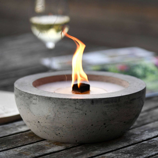 Detailansicht: Brennendes  Betonfeuer SAVO mit Durchmesser 25 cm, vor Weinglas  auf Holztisch.