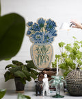 Blumenvasen-Teppich Blue Daisy an der Wand über einem Tisch mit Pflanzen, Kerzenhalter und Figuren