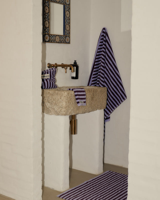 Kulturbeutel NARAM in Wine & Lavender Blue auf einem Waschbecken aus Naturstein, daneben Handtücher und Badematte im gleichen Streifenmuster