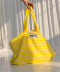 Hand hält die Weekend-Bag Pristine & Neon Yellow an den Henkeln vor einem weißen Vorhang im Sonnenlicht