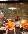 Zwei Gläser San Orangello mit Eis und Orangenscheibe, dazwischen die Flasche, daneben Baguette und eine Dose Sardinen