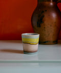 Kaffeebecher POP ART in glint mit gelbem Streifen zwischen Hellblau und Sand, vor roter Wand neben einem alten Steingutkrug