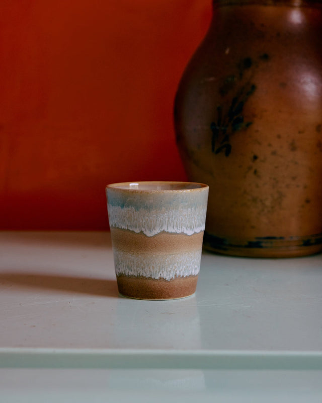 Kaffeebecher POP ART in skyline mit weißen Schlieren auf Sandbraun, vor roter Wand neben einem alten Steingutkrug