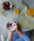 Gedeckter Tisch mit der KESÄKUKKA Leinen-Tischdecke: Beeren in einer Schale, ein Glas Orangensaft und eine Frau, die eine Schale Beeren mit Milch hält