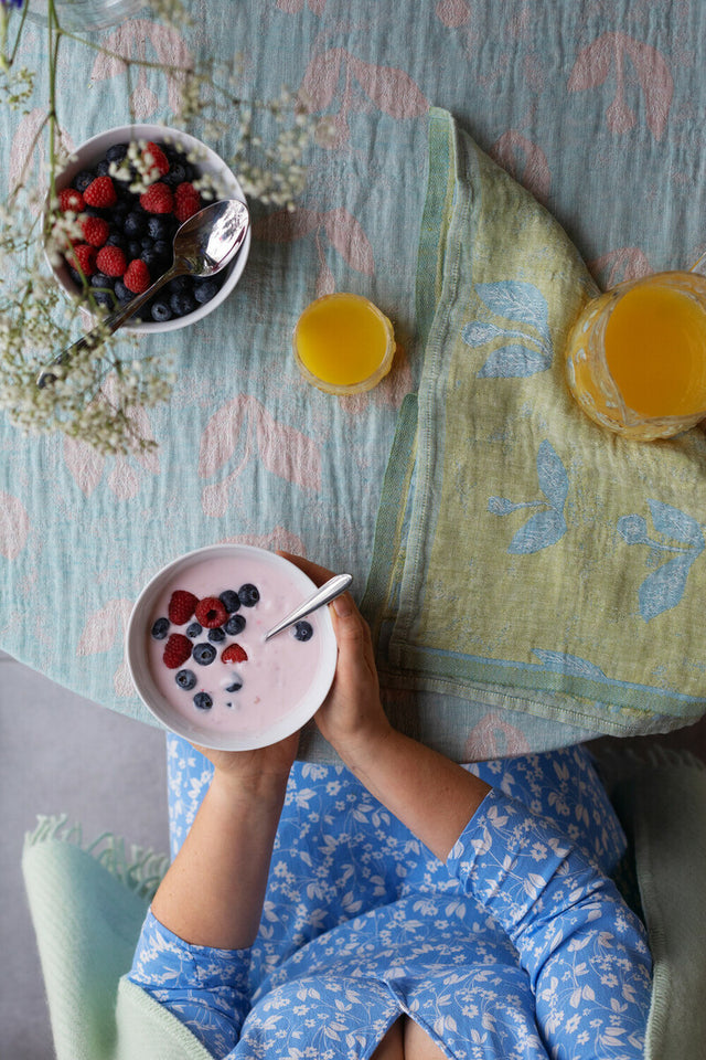 Gedeckter Tisch mit der KESÄKUKKA Leinen-Tischdecke: Beeren in einer Schale, ein Glas Orangensaft und eine Frau, die eine Schale Beeren mit Milch hält