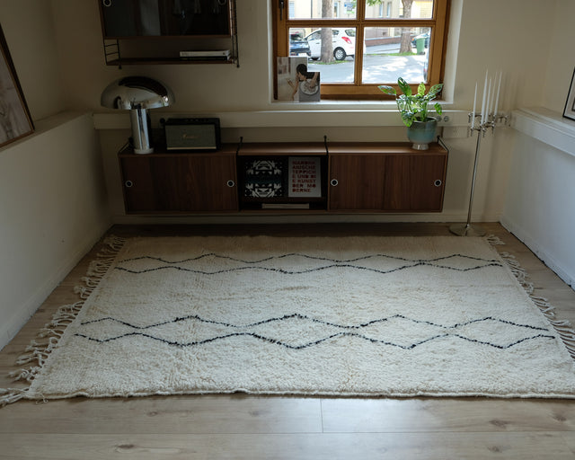 Der Azilal liegt in ganzer Fläche vor einem Sideboard aus dunklem Holz, schwarze Linien zeichnen ein Zickzackmuster auf den wollweißen Grund.
