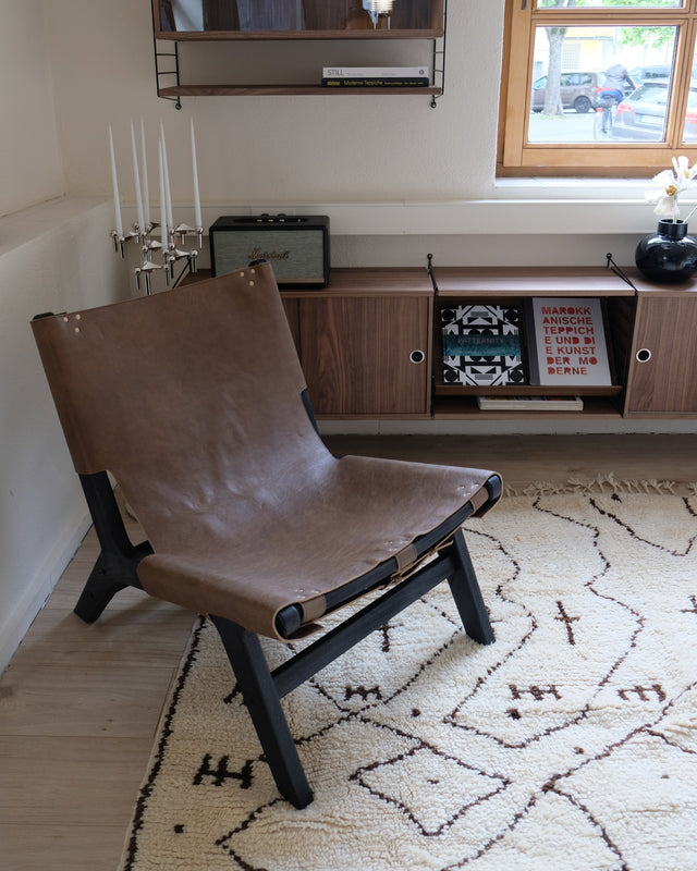 Wollweißer Berberteppich mit braunen Linien und Zeichen unter einem Ledersessel, dahinter ein Sideboard aus Holz