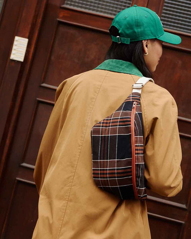 Person in beiger Jacke und grüner Kappe trägt die Bauchtasche BUM BAG in Black Tartan quer über dem Rücken