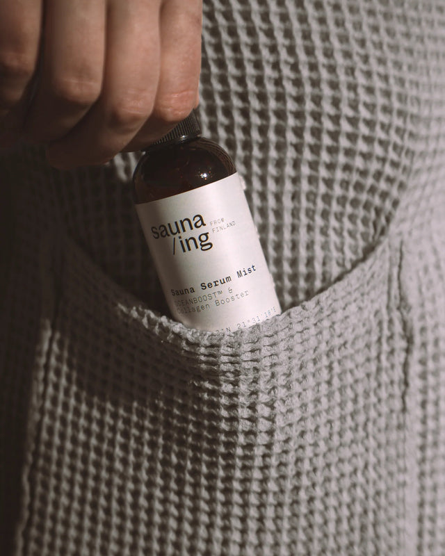 Hand steckt das Gesichtsspray Sauna Serum Mist in die Tasche eines grauen Waffelpiqué-Bademantels