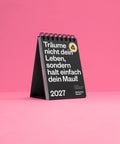 Schwarzer Tischkalender 2027 von Schlechte Karten vor rosa Hintergrund, aufgestellt mit der Titelseite nach vorn.