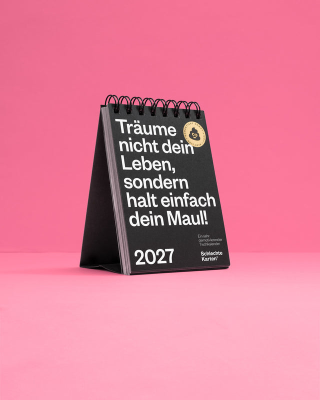 Schwarzer Tischkalender 2027 von Schlechte Karten vor rosa Hintergrund, aufgestellt mit der Titelseite nach vorn.