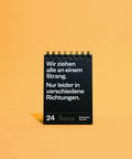 Tischkalender vor orangefarbenem Hintergrund, aufgeschlagen beim 24. Juni mit dem Spruch über den Strang in verschiedene Richtungen.