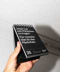Eine Hand hält den schwarzen Tischkalender vor eine weiße Wand, aufgeschlagen beim 25. Juni.