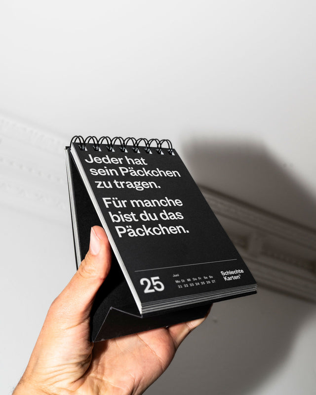 Eine Hand hält den schwarzen Tischkalender vor eine weiße Wand, aufgeschlagen beim 25. Juni.