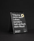 Titelseite des Tischkalenders 2027 mit goldrundem Aufkleber, fotografiert auf schwarzem Hintergrund.