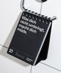 Der Tischkalender hängt an einem verchromten Handtuchhalter über weißen Badezimmerfliesen.