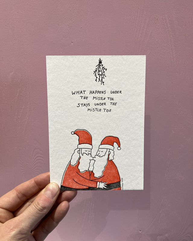 Letterpress-Weihnachtskarte von Studio Flash: zwei Weihnachtsmänner in roten Mänteln küssen sich unter einem Mistelzweig, darüber „What happens under the mistle toe stays under the mistle toe“, in der Hand vor einer rosa Holzwand