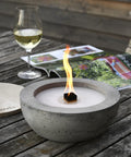 Brennendes  Betonfeuer SAVO mit Durchmesser 25 cm, vor Weinglas und Zeitschrift auf Holztisch.