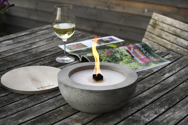 Brennendes  Betonfeuer SAVO mit Durchmesser 25 cm, vor Weinglas und Zeitschrift auf Holztisch.