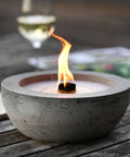 Detailansicht: Brennendes  Betonfeuer SAVO mit Durchmesser 25 cm, vor Weinglas  auf Holztisch.
