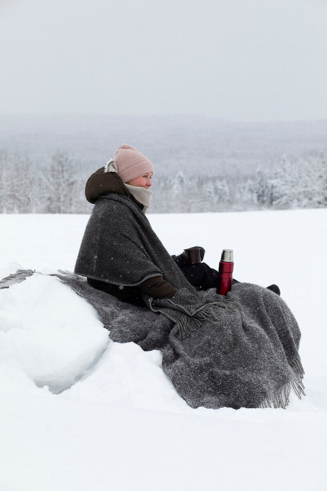 Wolldecke Maria mit Fischgrätmuster in Grau mit Fransen, umhüllt eine Frau beim Picknick im Schnee